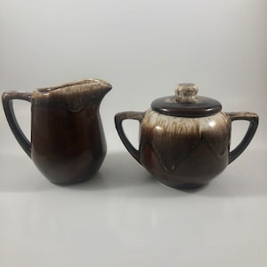 Puede incluir: Un juego de jarra de crema y azucarero de cerámica esmaltada en marrón y blanco. La jarra de crema tiene un solo asa y el azucarero tiene una tapa y dos asas.