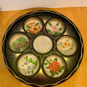 May include: A black metal tray with six circular compartments, each featuring a different floral design. The tray has a gold rim and decorative accents. The centre compartment has a white label with text.