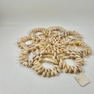 May include: A collection of white and brown shell necklaces, arranged in a circular pattern on a white background. The necklaces are made of small, oval-shaped shells, and there is a small white tag with the number "12" on it.