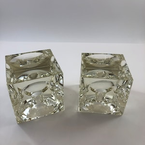 May include: A pair of clear glass candle holders with a square base and a round top. The candle holders are made of thick glass and have a textured surface.