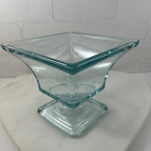 May include: A clear glass pedestal bowl with a square base and a square top. The bowl is made of clear glass and has a slight blue tint.