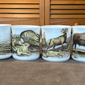 May include: Set of four white ceramic mugs with wildlife illustrations. Each mug features a different animal: a deer, a rabbit, a mountain goat, and a buffalo. The illustrations are in brown, black, and white.