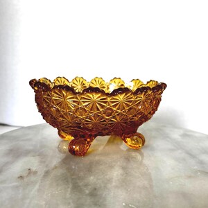 May include: An amber-colored glass bowl featuring a pressed starburst and diamond pattern across its exterior surface. The bowl has a scalloped rim and is supported by three small, curled feet, showcasing a translucent and reflective finish.