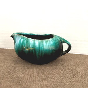 May include: A vintage, green ceramic creamer with a handle and spout. The creamer has a glossy finish with dark green and teal tones. The creamer is on a brown surface.
