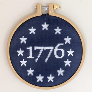 May include: A cross-stitch embroidery in a wooden hoop. The navy blue fabric features the year "1776" in white, surrounded by white stars. The hoop is tan and has a metal tightening mechanism.
