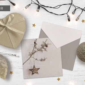 May include: A rustic Christmas card with a brown star hanging from a branch. The card is surrounded by a brown envelope, a heart-shaped box with a gold ribbon, a brown pine cone ornament, and a brown sphere ornament. The background is a white wooden surface with gold stars and a string of white lights.