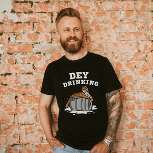 Puede incluir: Camiseta negra con el texto "DEY DRINKING" y un tigre de dibujos animados sobre un barril. El tigre es naranja y negro. El barril es gris. El fondo es una pared de ladrillos rojos.