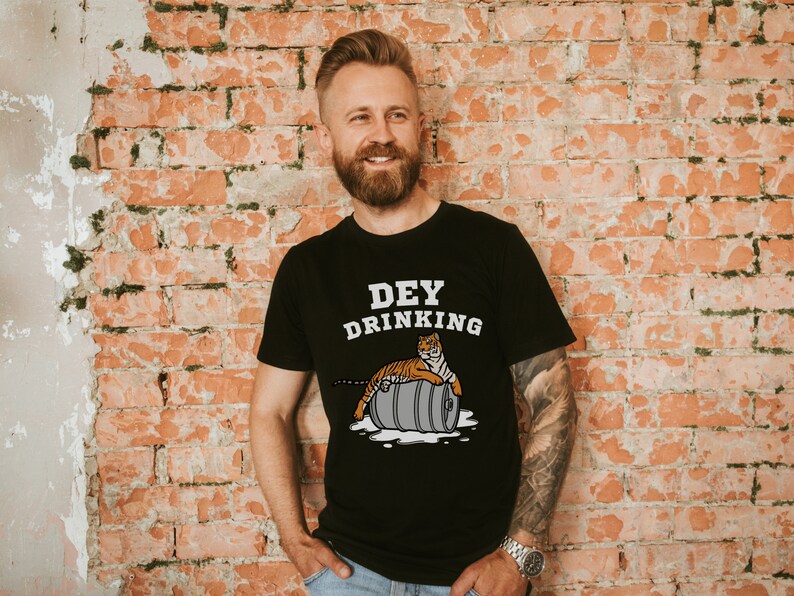 Puede incluir: Camiseta negra con el texto "DEY DRINKING" y un tigre de dibujos animados sobre un barril. El tigre es naranja y negro. El barril es gris. El fondo es una pared de ladrillos rojos.
