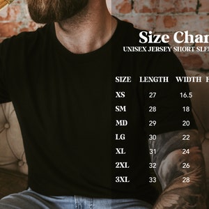 Puede incluir: Camiseta negra unisex de manga corta de punto de jersey con una tabla de tallas. La tabla de tallas enumera las medidas de XS a 3XL, incluyendo largo y ancho en pulgadas, y tallas correspondientes. La camiseta la lleva una persona.