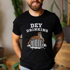 Puede incluir: Camiseta negra con el texto blanco "DEY DRINKING". Debajo, un dibujo de un tigre naranja y negro tumbado sobre un barril gris. El dise&ntilde;o est&aacute; impreso en la parte delantera de la camiseta.