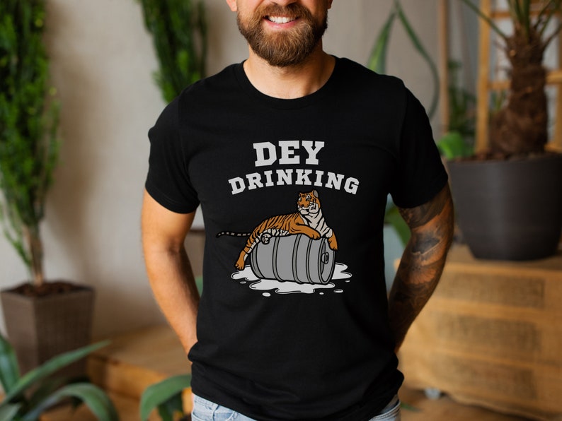 Puede incluir: Camiseta negra con el texto blanco "DEY DRINKING". Debajo, un dibujo de un tigre naranja y negro tumbado sobre un barril gris. El dise&ntilde;o est&aacute; impreso en la parte delantera de la camiseta.