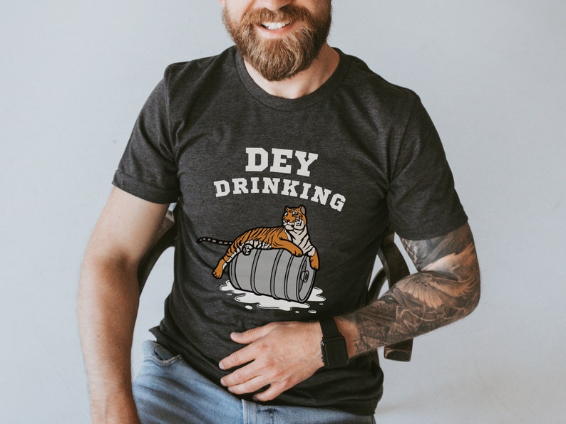 Puede incluir: Camiseta gris oscuro con el texto "DEY DRINKING" sobre una ilustraci&oacute;n de un tigre descansando sobre un barril de cerveza. El tigre es naranja y negro. La camiseta es de cuello redondo.