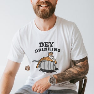Puede incluir: Camiseta blanca con el texto "DEY DRINKING" sobre un tigre de dibujos animados tumbado en un barril. El tigre es naranja y negro. El barril es gris. La camiseta es de manga corta.