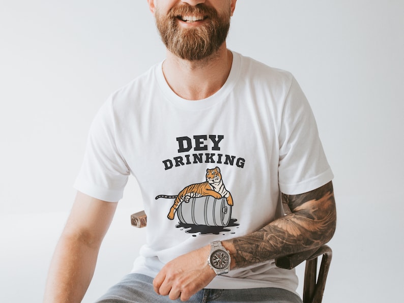 Puede incluir: Camiseta blanca con el texto "DEY DRINKING" sobre un tigre de dibujos animados tumbado en un barril. El tigre es naranja y negro. El barril es gris. La camiseta es de manga corta.