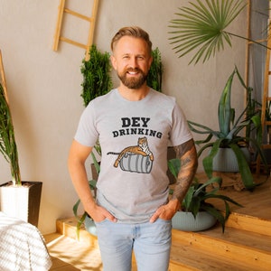 Puede incluir: Camiseta gris claro con el texto "Dey Drinking" y un dibujo de un tigre sobre un barril. El tigre es naranja y blanco. La camiseta la lleva una persona.