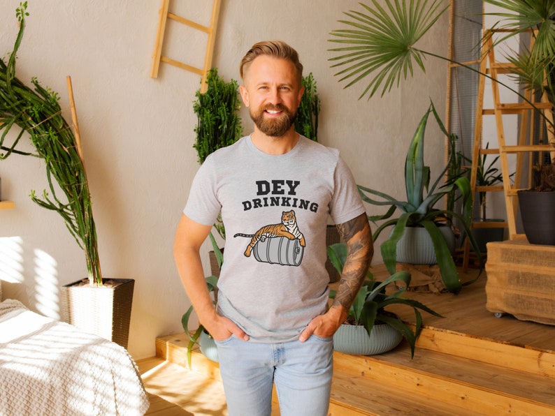 Puede incluir: Camiseta gris claro con el texto "Dey Drinking" y un dibujo de un tigre sobre un barril. El tigre es naranja y blanco. La camiseta la lleva una persona.