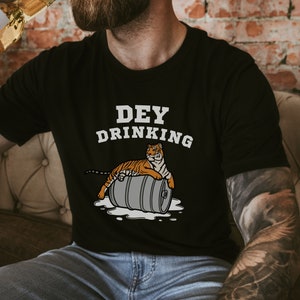 Puede incluir: Camiseta negra con el texto "DEY DRINKING" y un tigre de dibujos animados sobre un barril. El tigre es naranja y negro. La camiseta la lleva una persona que sostiene una botella.