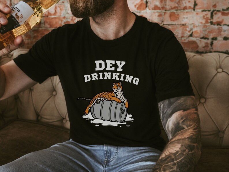 Puede incluir: Camiseta negra con el texto "DEY DRINKING" y un tigre de dibujos animados sobre un barril. El tigre es naranja y negro. La camiseta la lleva una persona que sostiene una botella.
