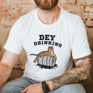 Puede incluir: Camiseta blanca con el texto "DEY DRINKING" y un tigre de dibujos animados descansando sobre un barril. El tigre es naranja y negro. El barril es gris con l&iacute;quido negro saliendo.