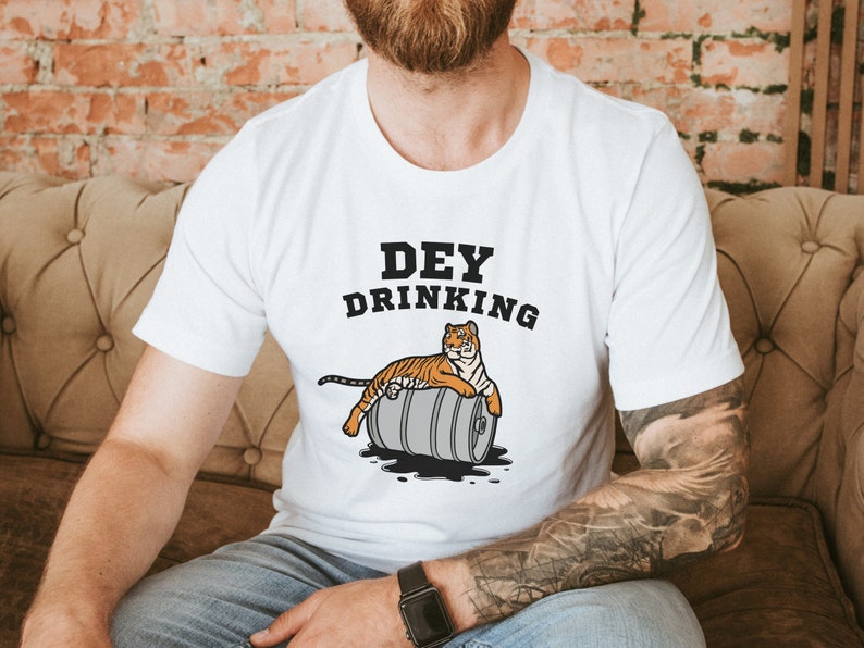 Puede incluir: Camiseta blanca con el texto "DEY DRINKING" y un tigre de dibujos animados descansando sobre un barril. El tigre es naranja y negro. El barril es gris con l&iacute;quido negro saliendo.