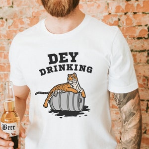 Puede incluir: Camiseta blanca con el texto "Dey Drinking" sobre un tigre de dibujos animados descansando sobre un barril. El tigre es naranja y negro. Una botella de cerveza se sostiene en la mano de la persona.