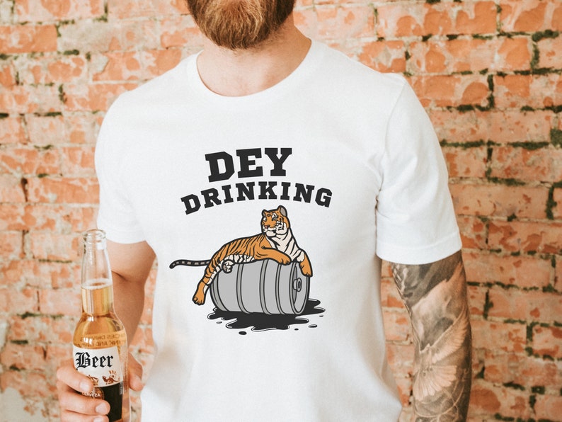Puede incluir: Camiseta blanca con el texto "Dey Drinking" sobre un tigre de dibujos animados descansando sobre un barril. El tigre es naranja y negro. Una botella de cerveza se sostiene en la mano de la persona.