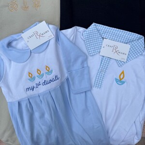 May include: Two baby outfits, one light blue and white with a round collar and the text "my 1st diwali". The other is white with a blue checkered collar. Both have a tag that says "Crafty Mama". Each outfit has an embroidered diya design.