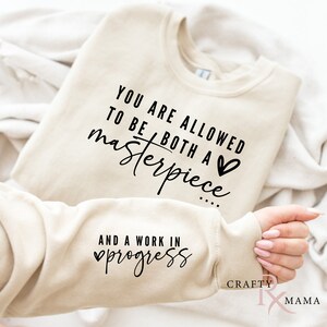 以下が含まれることがあります： ベージュのトレーナーに「YOU ARE ALLOWED TO BE BOTH A masterpiece... AND A WORK IN progress」という黒い文字がプリントされています。