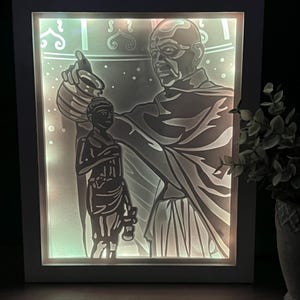 May include: A white framed light-up art piece depicting a silhouette of a person pouring water from a pot onto a smaller person. The background is a light blue with white dots.