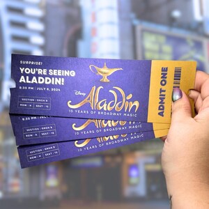 Puede incluir: Un montón de entradas moradas y doradas para el espectáculo de Broadway Aladdin. Las entradas dicen "¡Sorpresa! ¡Vas a ver Aladdin!" y "10 años de magia en Broadway".