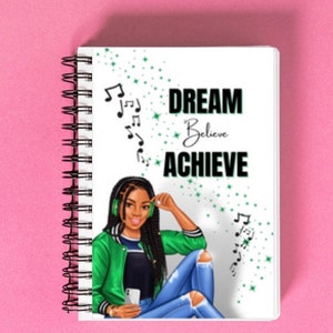 May include: A white spiral bound notebook with a green and black illustration of a woman wearing headphones and a green jacket. The notebook has the text "DREAM Believe ACHIEVE" on the front.