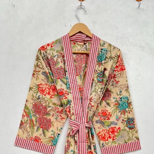 Puede incluir: Bata con estampado floral beige con detalles en rojo y azul. La bata presenta un ribete y cinturón a rayas. La bata está colgada de una percha de madera contra una pared blanca.