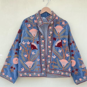 Puede incluir: Chaqueta bordada azul claro con diseño floral. La chaqueta tiene cuello alto, mangas largas y forro a rayas. El bordado incluye flores y hojas rosas, rojas y naranjas. La chaqueta está colgada de una percha de madera.