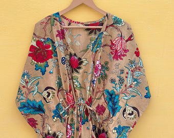 Peut inclure: Robe caftan beige à imprimé floral, col en V et taille à cordon. La robe présente un motif vibrant de fleurs et de feuillages rouges, bleus et roses. Elle est suspendue à un cintre en bois sur un fond jaune.