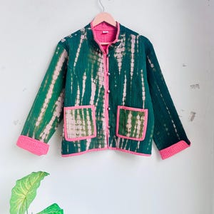 May include: A quilted jacket featuring a dark green and white tie-dye pattern with bright pink trim along the collar, front opening, cuffs, and two square front pockets. The jacket has a mandarin-style collar and a button-down front closure.