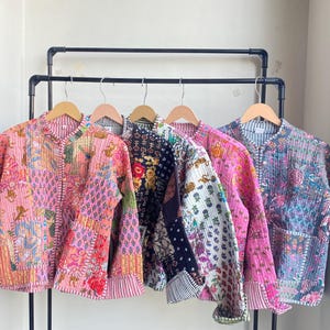Puede incluir: Cinco chaquetas acolchadas de colores cuelgan de un perchero de metal negro. Las chaquetas presentan una variedad de patrones y colores, incluyendo rosa, negro y azul. Cada chaqueta tiene un diseño de patchwork único y se exhibe en una percha de madera.