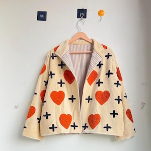 Puede incluir: Chaqueta beige con aplicaciones de corazón rojo y cruz negra. La chaqueta tiene un forro a rayas y un cuello. Cuelga de una percha de madera contra una pared blanca. La chaqueta es una prenda única.