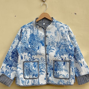 May include: A blue and white quilted jacket with a mandarin collar and two patch pockets hangs on a wooden hanger against a beige wall. The jacket features a toile-style print of birds and trees. The cuffs and pocket trim are in a navy and white striped fabric.  The jacket is lightweight and appears to be made of cotton. It is a casual style, suitable for spring or fall.