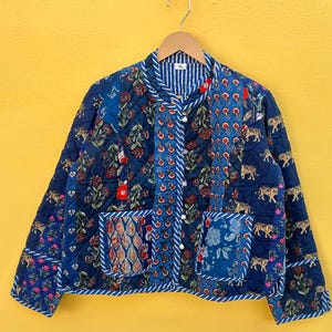 Op de afbeelding: Een blauw gewatteerd jack met een patchwork-ontwerp, met bloemen-, paisley- en tijgerpatronen. Het heeft een knoopsluiting, twee zakken en een gestreepte kraag. De jas hangt aan een houten hanger.