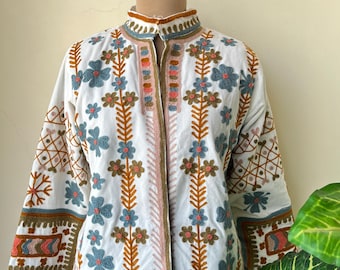 May include: White jacket with intricate floral embroidery in blue, brown, and pink. The jacket features a stand-up collar and detailed patterns on the sleeves. The embroidery creates a bohemian aesthetic, perfect for adding a unique touch to any outfit.