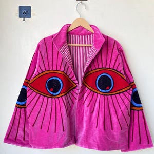 Puede incluir: Chaqueta de terciopelo rosa brillante con grandes diseños de ojos bordados en rojo, azul y naranja. La chaqueta tiene cuello y forro interior a rayas. Las mangas tienen detalles adicionales de ojos.