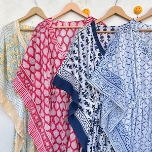 May include: Four colorful, patterned caftans hanging on wooden hangers. The caftans feature various designs, including leaf and geometric patterns, in shades of yellow, pink, white, and blue. The caftans are made of lightweight fabric.