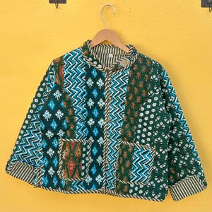 以下が含まれることがあります： 緑、ティール、茶色のパッチワークデザインのキルティングジャケット。 ジャケットはボタン留め、2つのポケット、ストライプの裏地が付いています。 木のハンガーに吊るされており、黄色の壁に掛けられています。