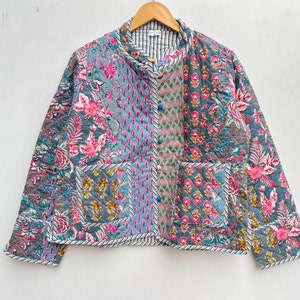 Puede incluir: Chaqueta acolchada con diseño patchwork. La chaqueta presenta una mezcla de estampados florales y geométricos en tonos grises, rosas y azules. Tiene cuello redondo, dos bolsillos y se abrocha con botones en la parte delantera. La chaqueta se muestra en una percha de madera.