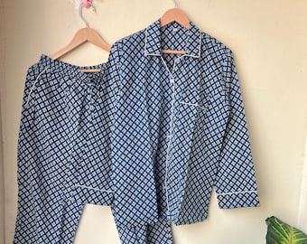 May include: A navy blue pajama set with a white geometric pattern. The set includes a button-down shirt with white piping and matching pants. The pajamas are hanging on wooden hangers against a neutral wall.
