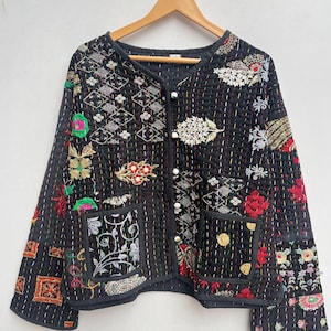 May include: Black quilted jacket with colourful floral and geometric embroidery. The jacket has a round neckline, long sleeves, and two front pockets. It is adorned with white buttons and hangs on a wooden hanger.