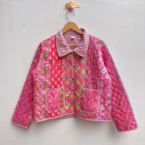 Puede incluir: Chaqueta acolchada rosa con estampados florales y geométricos. La chaqueta tiene cuello y mangas largas. El diseño incluye patchwork y costuras acolchadas, con forro y ribete a rayas. La chaqueta está colgada en una percha de madera.