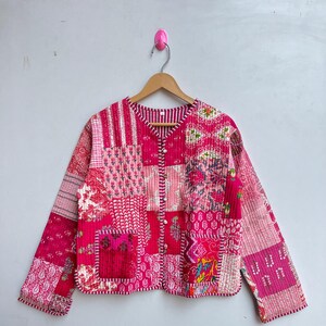 Può includere: Una giacca trapuntata rosa con un design a patchwork. La giacca ha un collo a V e maniche lunghe. Il tessuto è un mix di diversi motivi e colori, tra cui rosa, rosso e bianco.