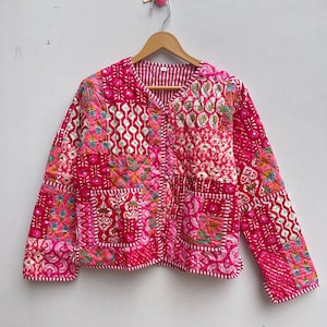 May include: A pink quilted jacket with a patchwork design, showcasing floral and geometric patterns. The jacket has a V-neck, long sleeves, and two front pockets. The lining features a striped pattern.