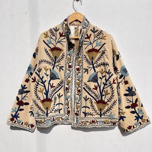 Puede incluir: Una chaqueta beige bordada con un patrón floral en tonos azules, rojos y dorados. La chaqueta tiene un cuello alto y mangas largas.